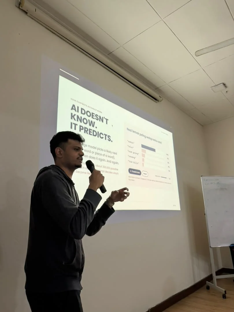 An OrynLab trainer with a microphone in front of a slide reading 'AI doesn't know. It predicts.'